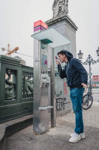 man wearing blue jeans and jacket shouting at a telephone booth 