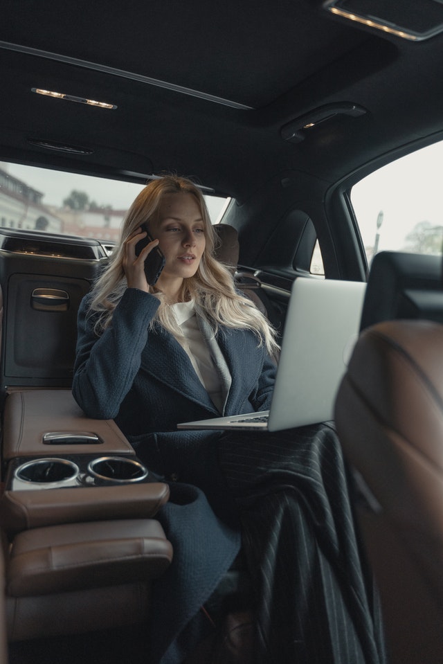 a woman in corporate attire answering a call in her car with laptop on her lap